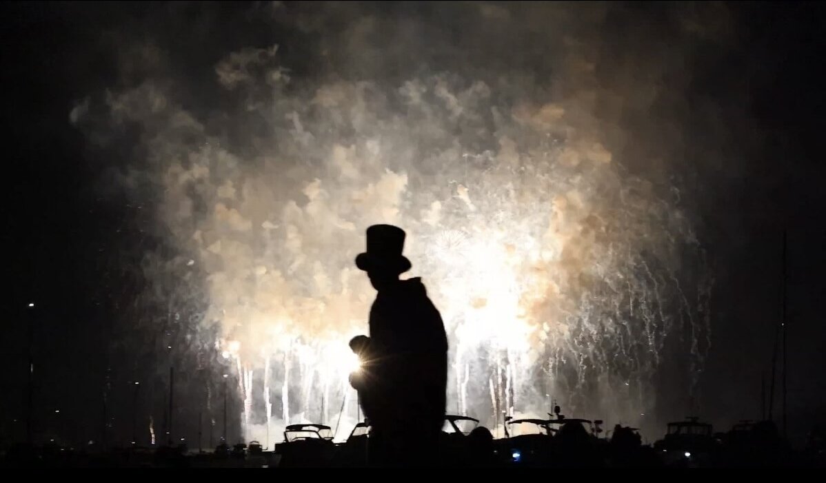 A top-hatted statue silhouetted against fireworks and smoke